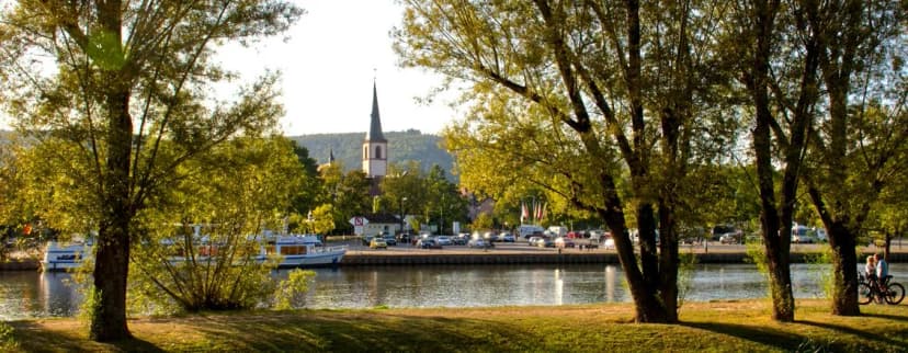Blick über den Main auf Lohr mit Kirchturm, Booten am Ufer und Bäumen im Vordergrund