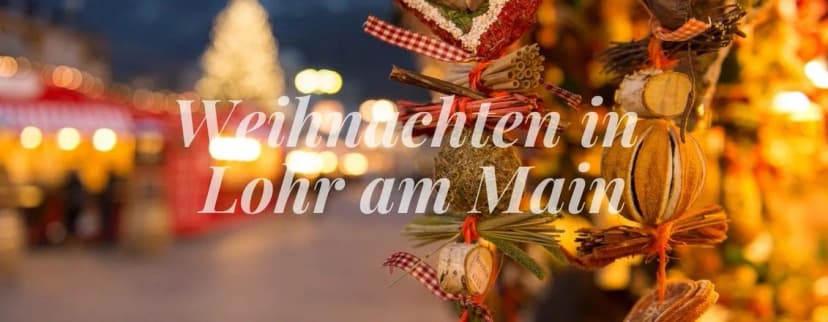 Weihnachtlicher Marktstand mit Dekoration aus Zweigen und Bändern, im Hintergrund beleuchteter Weihnachtsmarkt mit Christbaum, Schriftzug Weihnachten in Lohr am