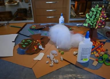 Bastelmaterial für ein Landschaftsmodell: Klebstoffflaschen, Watte, Papierbäume mit bunten Blättern und ausgeschnittene braune Papierformen auf einem Tisch.