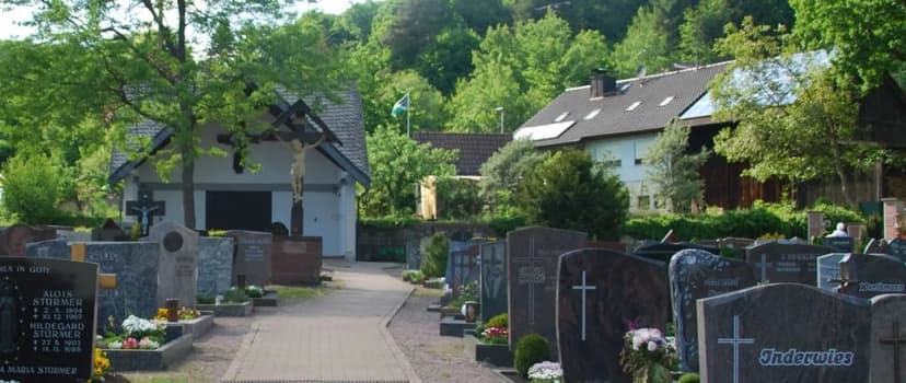 Friedhof mit Grabsteinen, Leichenhalle mit Kruzifix und Wohnhäusern im Hintergrund, umgeben von bewaldetem Hang