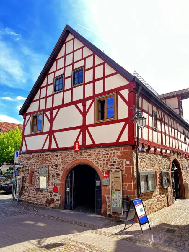 Fachwerkhaus mit rot-weißem Gebälk und Sandsteinsockel, Eingang zur Touristinformation mit Infoschild
