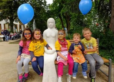 Kinder sitzen mit blauen Luftballons auf einer Bank neben einer steinernen Frauenfigur im Park