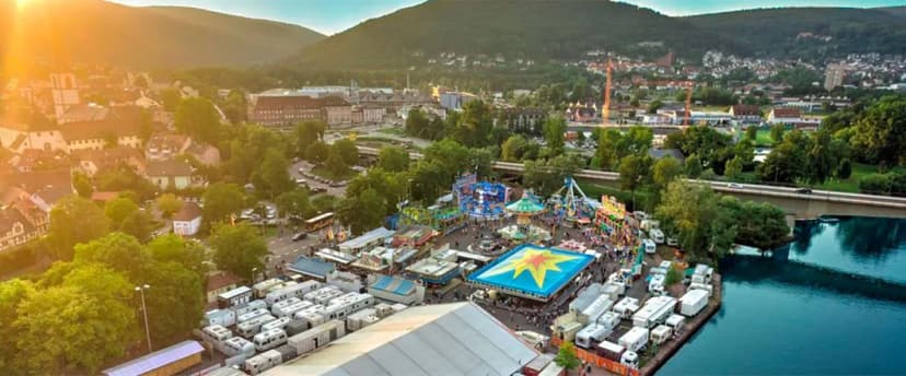 Luftaufnahme eines Volksfestgeländes mit Fahrgeschäften am Flussufer, umgeben von Stadt und bewaldeten Hügeln.