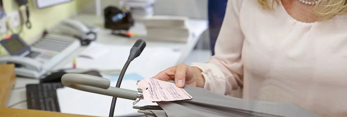 Person hält an einem Schreibtisch mit Mikrofon und Telefon einen handschriftlich ausgefüllten Zettel über einem Kleidungsstück