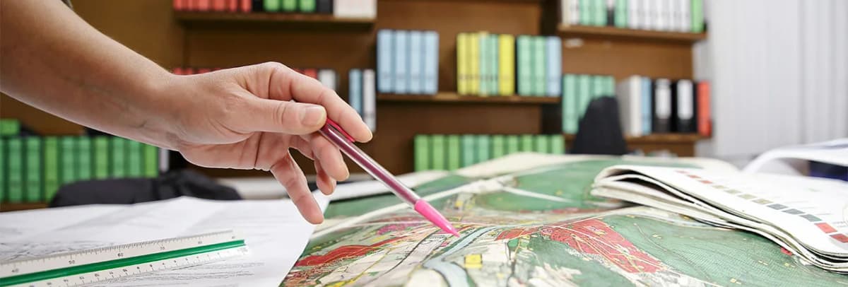 Hand mit Stift zeigt auf eine historische Landkarte, im Hintergrund ein Bücherregal