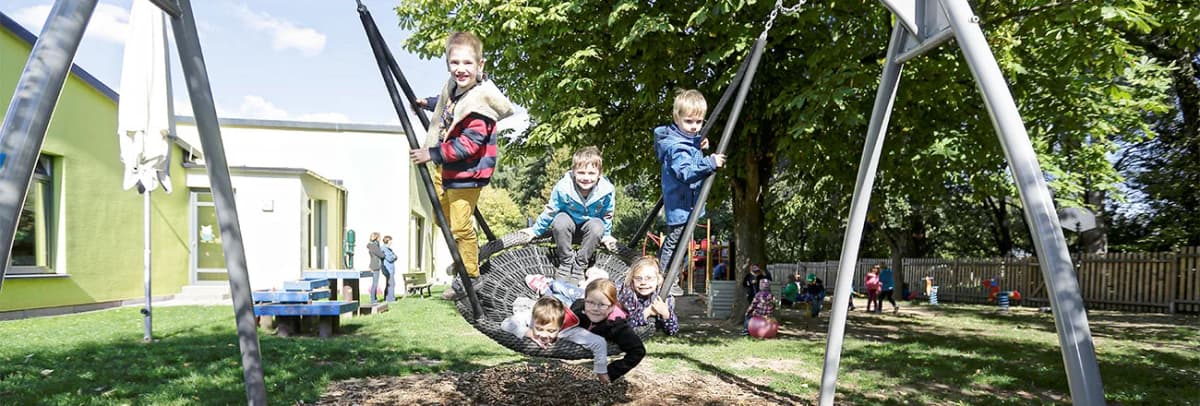 Kinder spielen auf einer Nestschaukel im Freien vor einem Kindergartengebäude mit Garten und Spielplatz