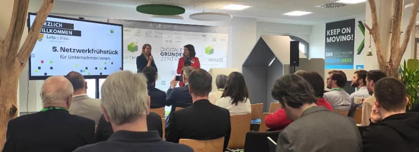 Zwei Frauen sprechen vor Publikum im Digitalen Gründerzentrum, Bildschirm zeigt Willkommensgruß zum 5. Netzwerkfrühstück für Unternehmerinnen