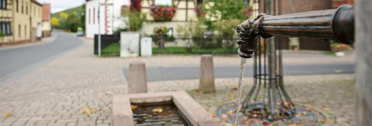 Dorfbrunnen mit fließendem Wasser, im Hintergrund Fachwerkhäuser und eine Straße