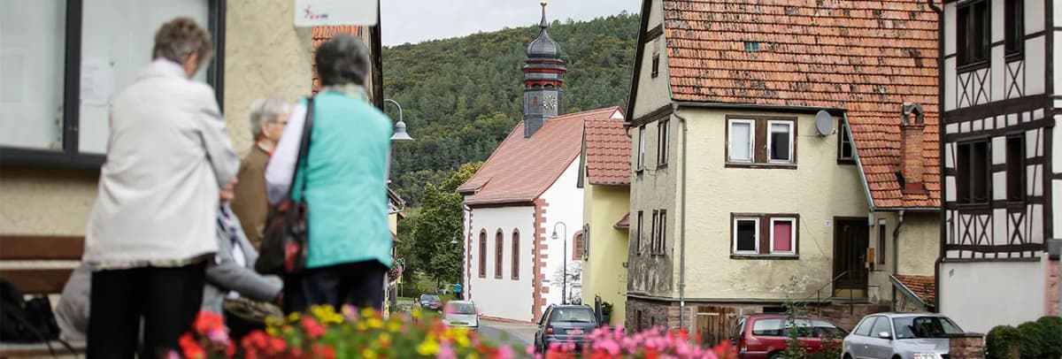 Passanten in einer Ortsstraße mit Kapelle, Fachwerkhäusern und bewaldetem Hügel im Hintergrund