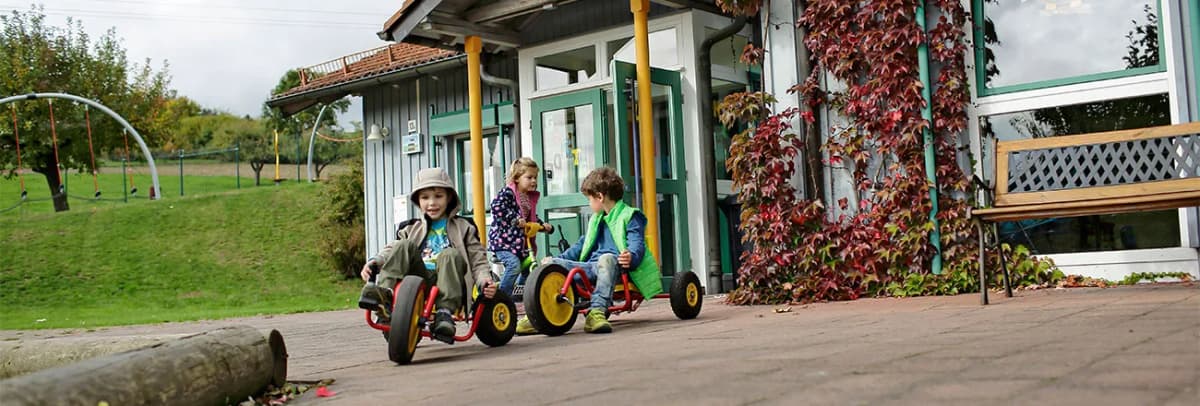 Kinder fahren mit Dreirädern vor einem Kindergartengebäude mit Spielplatz und Rankpflanzen an der Fassade
