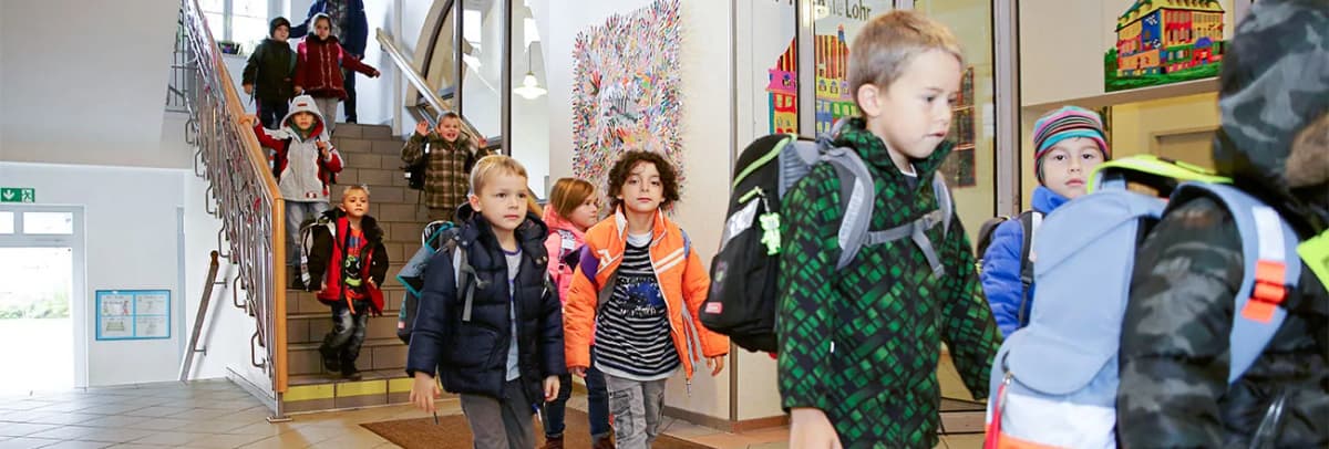 Kinder mit Schulranzen gehen im Schulflur eine Treppe herunter und durch das Foyer.