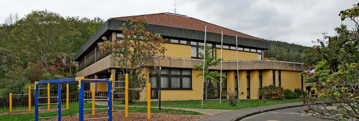 Gelbes zweistöckiges Gebäude mit Walmdach und davorliegendem Spielplatz mit Klettergerüst