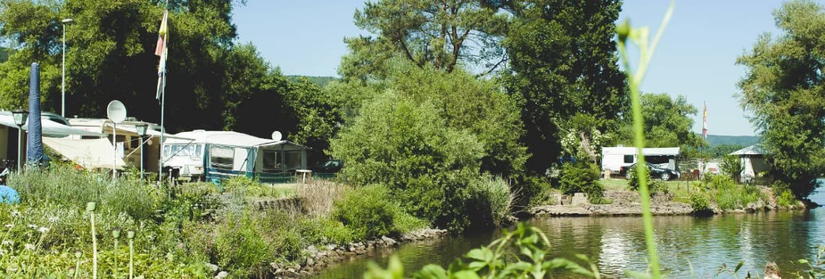 Campingplatz mit Wohnwagen am Flussufer, umgeben von Bäumen und Grünflächen.