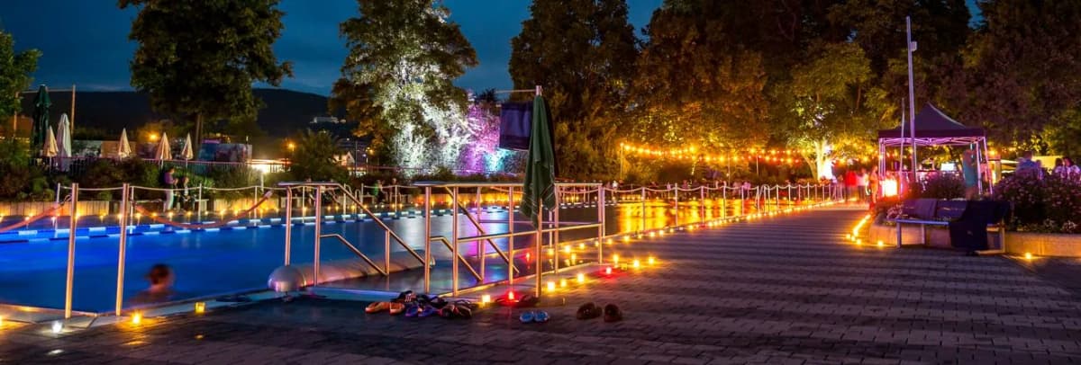 Freibad bei Nacht mit Kerzen entlang des Beckenrands, Lichterketten und beleuchteten Bäumen während einer Candle-Light-Veranstaltung.