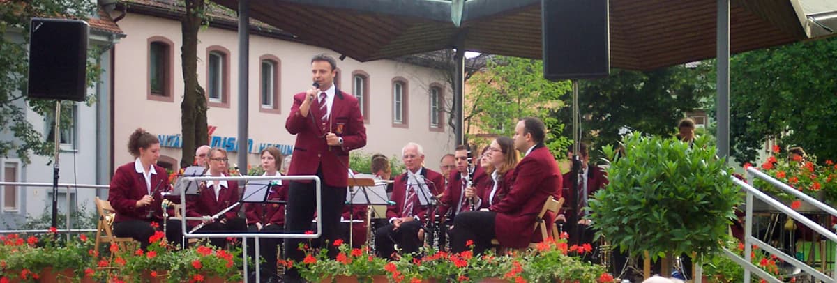 Musikkapelle in bordeauxroten Uniformen tritt mit Blasinstrumenten auf einer mit Blumenkästen geschmückten Freilichtbühne auf, ein Sänger spricht ins Mikrofon.
