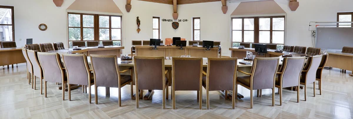 Sitzungssaal mit hufeisenförmig angeordneten Tischen, Stühlen und Bildschirmen für die Ratsmitglieder