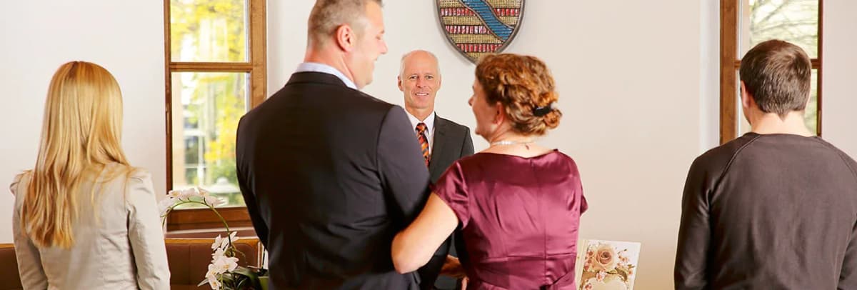 Trauung im Standesamt: Ein Mann in Anzug begrüßt ein Paar vor einem Wappen an der Wand, Gäste stehen dabei.