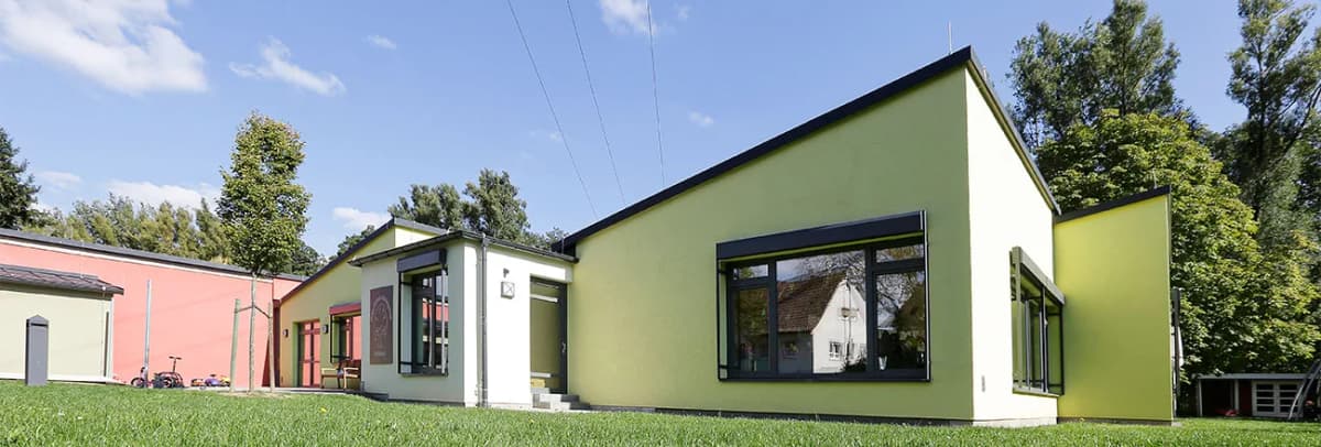 Eingeschossiges Kindergarten- oder Schulgebäude mit gelb-grüner und roter Fassade, großen Fenstern und Rasenfläche davor