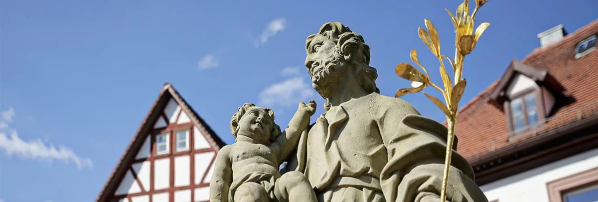 Steinerne Heiligenfigur mit Kind und goldenem Lilienzweig vor Fachwerkhäusern