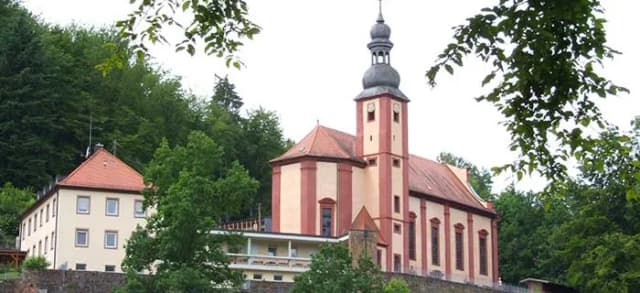 Kirche mit Turm und Zwiebelhaube neben Klostergebäude, umgeben von bewaldetem Hang
