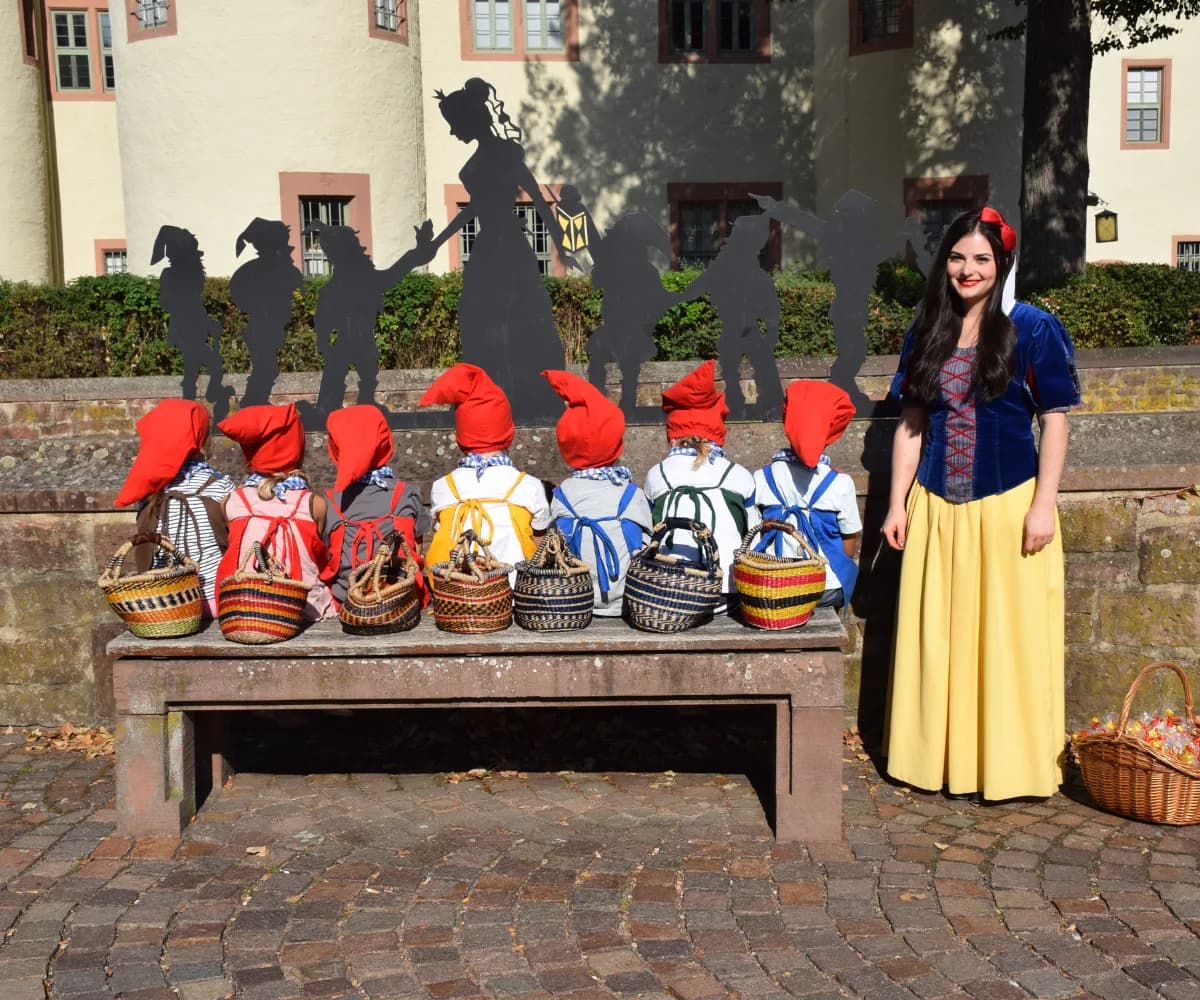 Schneewittchen und ihre sieben Zwerge, die jeweils rote Mützen tragen und mit dem Rücken zum Betrachter auf einer Bank sitzen, sind vor dem Schloss in Lohr am Main zu sehen. Dahinter befindet sich die Schneewittchen-Silhouette.