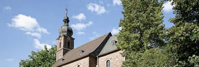 Kirchturm mit Zwiebelhaube neben Kirchendach, umgeben von Bäumen vor blauem Himmel