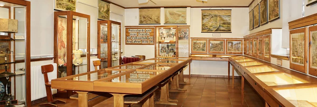 Ausstellungsraum eines Schulmuseums mit Glasvitrinen, historischen Schautafeln und Bildern an den Wänden.