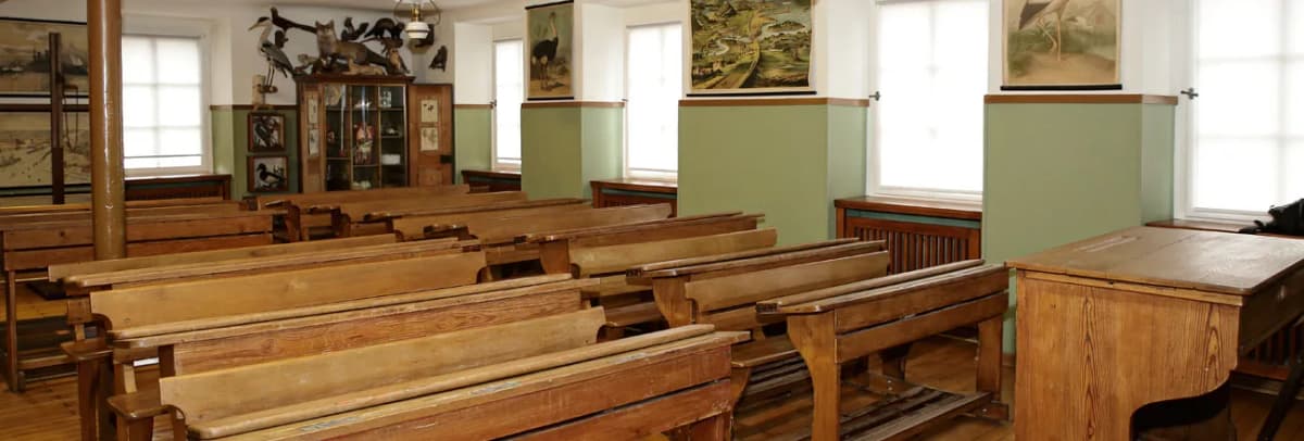 Historisches Klassenzimmer mit alten Holzbänken, Lehrerpult und Wandbildern von Vögeln zwischen den Fenstern