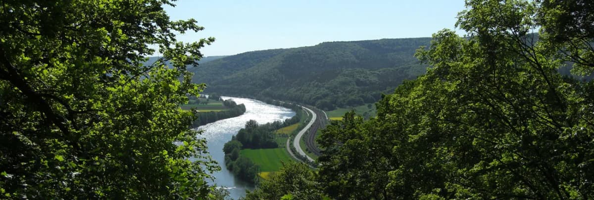 Blick durch Bäume auf ein Flusstal mit Bahnstrecke und bewaldeten Hügeln